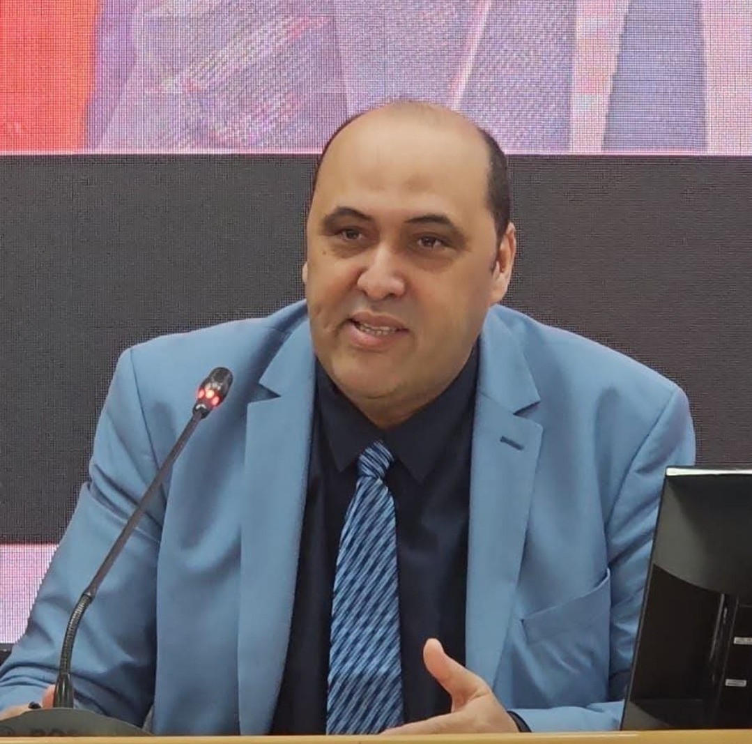 WhatsApp Image 2026-06-22 at 120859 | MCG24 | موقع إلكتروني مغربي شامل متجدد على مدار الساعة Man in a light-blue suit and striped tie speaks into a microphone at a conference podium, gesturing with his hand.