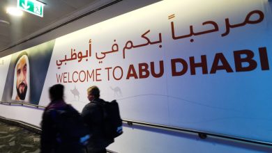 Large bilingual welcome sign on a curved wall reading 'WELCOME TO ABU DHABI' with Arabic text above; people walk past in a corridor along a handrail.