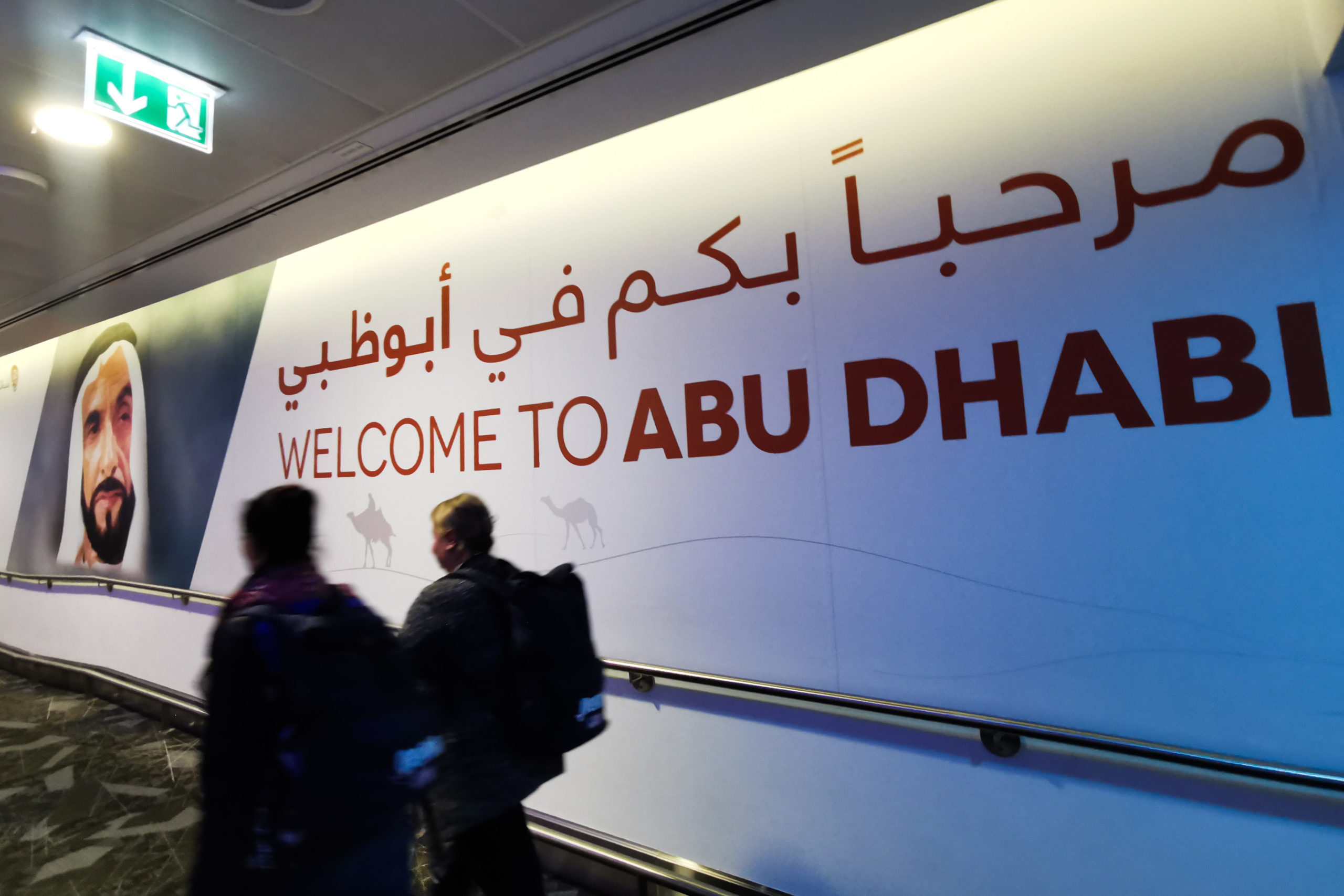 Large bilingual welcome sign on a curved wall reading 'WELCOME TO ABU DHABI' with Arabic text above; people walk past in a corridor along a handrail.