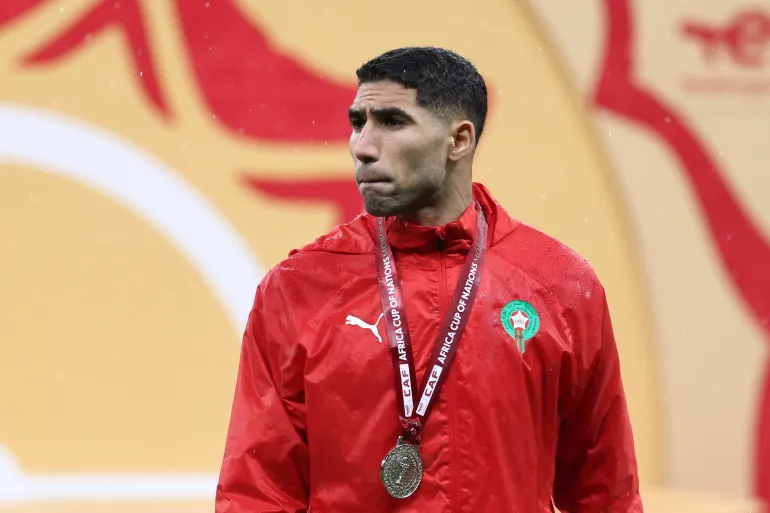 Man in a red track jacket with a medal on a lanyard around his neck, standing at a sports event background.
