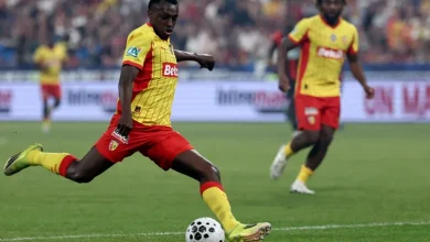 Soccer player in a yellow and red kit kicking a ball on a green field, with a teammate blurred in the background at a crowded stadium