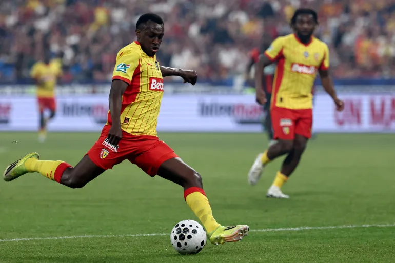 Soccer player in a yellow and red kit kicking a ball on a green field, with a teammate blurred in the background at a crowded stadium