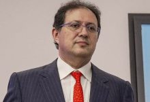 ait-mena-2 | MCG24 | موقع إلكتروني مغربي شامل متجدد على مدار الساعة Man in a navy pinstripe suit with a red tie and glasses, standing indoors.