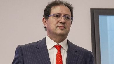 Man in a navy pinstripe suit with a red tie and glasses, standing indoors.