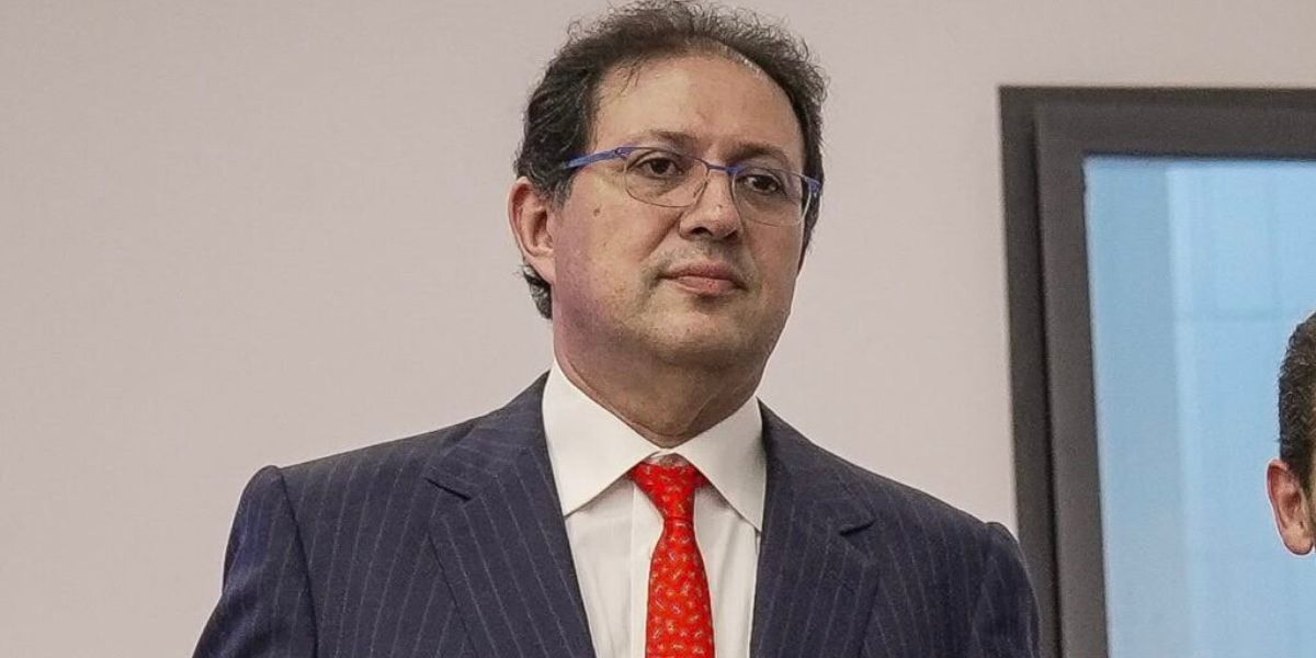ait-mena-2 | MCG24 | موقع إلكتروني مغربي شامل متجدد على مدار الساعة Man in a navy pinstripe suit with a red tie and glasses, standing indoors.