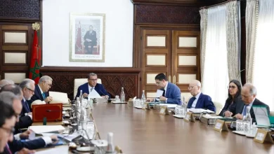 Group of professionals in suits seated around a long wooden conference table, with documents and glasses, in a room with wood paneling and a framed portrait on the wall.
