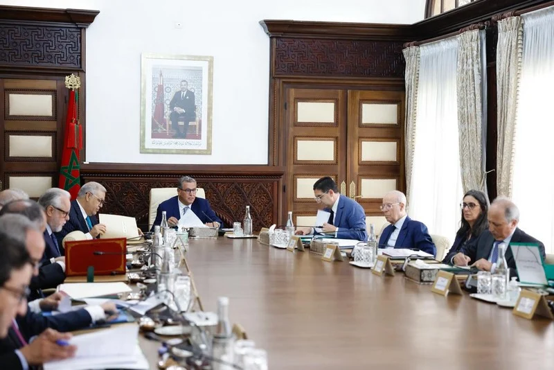 Group of professionals in suits seated around a long wooden conference table, with documents and glasses, in a room with wood paneling and a framed portrait on the wall.