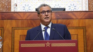 akhnnouch-86 | MCG24 | موقع إلكتروني مغربي شامل متجدد على مدار الساعة Official in a navy suit and blue tie speaks at a red podium with decorative gold trim, in a wood-paneled chamber with two microphones present.