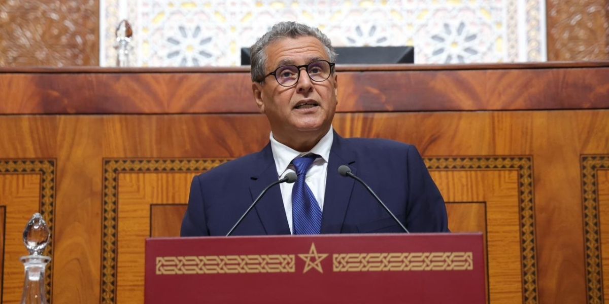 akhnnouch-86 | MCG24 | موقع إلكتروني مغربي شامل متجدد على مدار الساعة Official in a navy suit and blue tie speaks at a red podium with decorative gold trim, in a wood-paneled chamber with two microphones present.