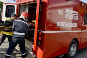 Paramedics load a patient on a stretcher into the back of a red ambulance labeled 'AMBULANCE' and 'PROTECTION CIVILE' (emergency scene)