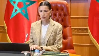 amira_1781284050 | MCG24 | موقع إلكتروني مغربي شامل متجدد على مدار الساعة A woman in a beige blazer seated at a desk, with Moroccan flags in the background, speaking at a formal meeting.