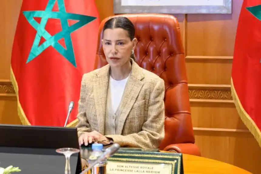 amira_1781284050 | MCG24 | موقع إلكتروني مغربي شامل متجدد على مدار الساعة A woman in a beige blazer seated at a desk, with Moroccan flags in the background, speaking at a formal meeting.
