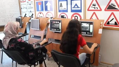 auto-ecole | MCG24 | موقع إلكتروني مغربي شامل متجدد على مدار الساعة Two learners sit at driving-simulation desks with screens in a classroom lined with road-sign posters behind them.
