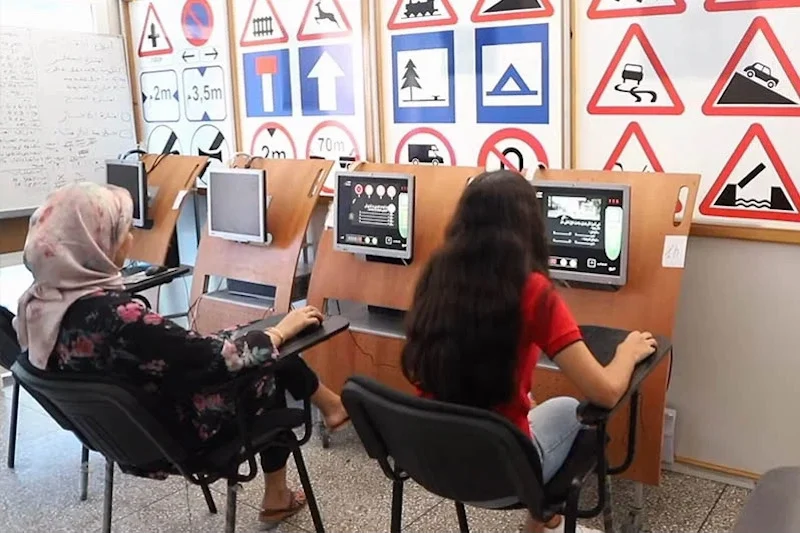 auto-ecole | MCG24 | موقع إلكتروني مغربي شامل متجدد على مدار الساعة Two learners sit at driving-simulation desks with screens in a classroom lined with road-sign posters behind them.