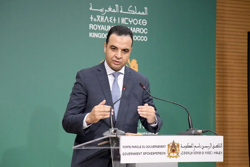 Government spokesperson in a dark suit speaking at a lectern with the Moroccan crest and signage behind him.