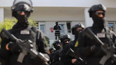 bcij-maroc | MCG24 | موقع إلكتروني مغربي شامل متجدد على مدار الساعة Heavy-armed tactical team in black gear stands outside a white building, with an officer on the balcony above.