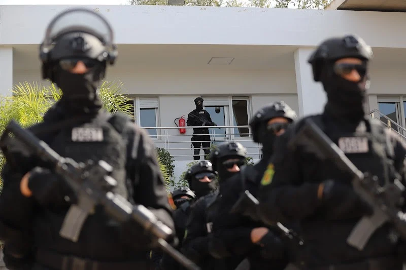 bcij-maroc | MCG24 | موقع إلكتروني مغربي شامل متجدد على مدار الساعة Heavy-armed tactical team in black gear stands outside a white building, with an officer on the balcony above.
