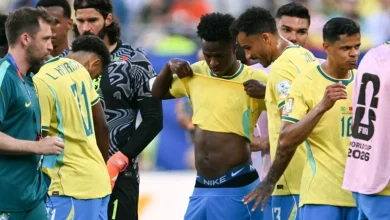 brazil-mondiale-2026-1 | MCG24 | موقع إلكتروني مغربي شامل متجدد على مدار الساعة Brazilian soccer players in yellow jerseys gather on the field in a team huddle, with one lifting his shirt to adjust it and a goalkeeper in black behind them. A pink bib on the right shows World Cup 2026 branding.