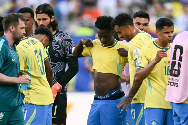 Brazilian soccer players in yellow jerseys gather on the field in a team huddle, with one lifting his shirt to adjust it and a goalkeeper in black behind them. A pink bib on the right shows World Cup 2026 branding.