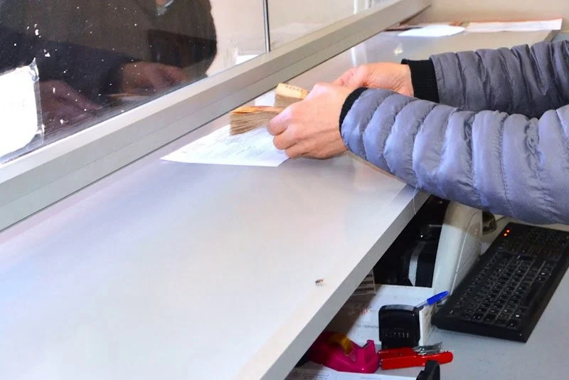 Person in a blue jacket wipes or brushes a document on a service counter beneath a glass divider at a reception or teller desk.