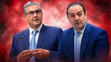 chaouki-1-585x389 | MCG24 | موقع إلكتروني مغربي شامل متجدد على مدار الساعة Two men in dark blue suits stand at a podium with a microphone against a red smoky background, speaking to an audience.