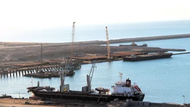 Cargo ship docked at a harbor with cranes along the pier and calm blue water nearby.