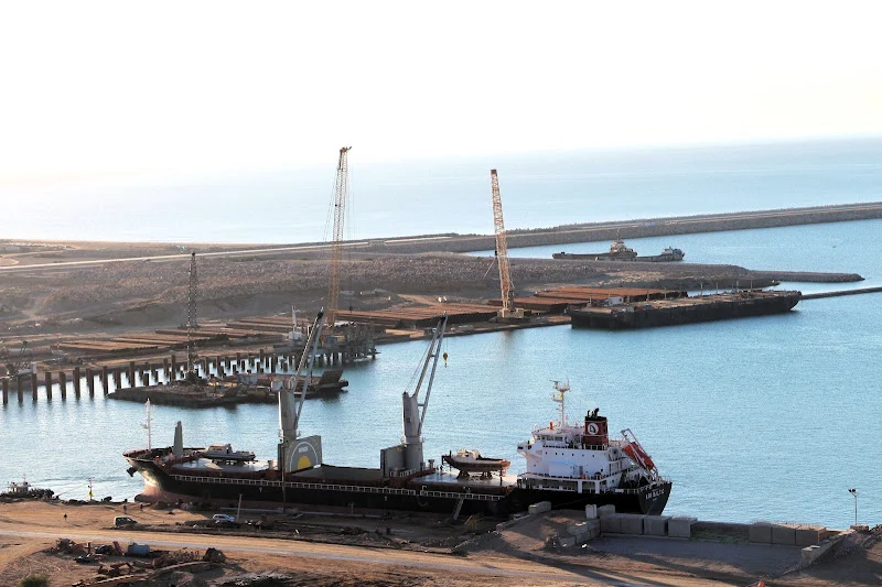 Cargo ship docked at a harbor with cranes along the pier and calm blue water nearby.
