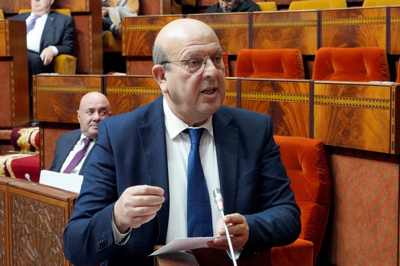 عبد الجبار الرشيدي | MCG24 | موقع إلكتروني مغربي شامل متجدد على مدار الساعة Middle-aged man in a suit and glasses speaking into a microphone in a parlament-like chamber, holding papers.