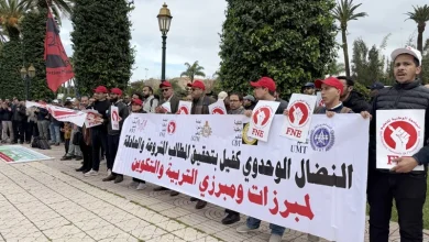 enseignants-sit-in | MCG24 | موقع إلكتروني مغربي شامل متجدد على مدار الساعة A group of protesters holding a long white banner with Arabic text, standing in a line outdoors among palm trees and lampposts, many wearing red caps and holding circular protest signs with a red fist logo.