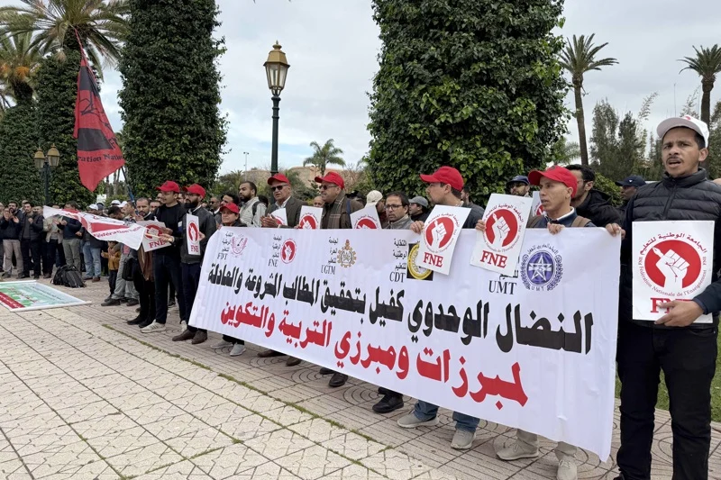 A group of protesters holding a long white banner with Arabic text, standing in a line outdoors among palm trees and lampposts, many wearing red caps and holding circular protest signs with a red fist logo.