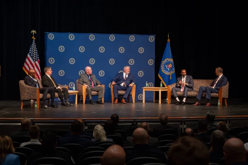 fifa-FBI | MCG24 | موقع إلكتروني مغربي شامل متجدد على مدار الساعة Six men in suits sit on stage in a panel discussion, with U.S. flag and blue emblem backdrop behind them; audience in foreground.