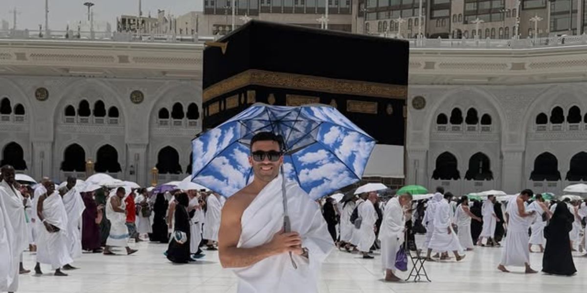 hamdallah | MCG24 | موقع إلكتروني مغربي شامل متجدد على مدار الساعة Smiling man in white ihram holding a blue sky umbrella in the courtyard of the Kaaba, with pilgrims around.