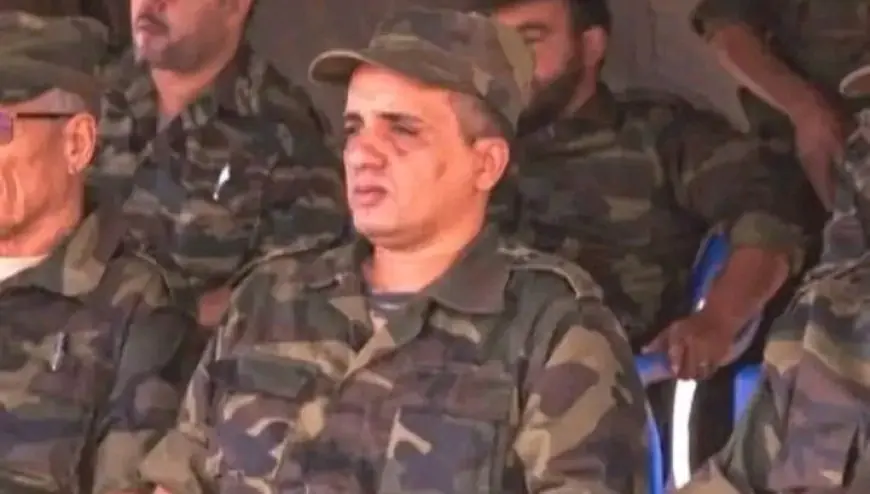 Group of soldiers in camouflage uniforms seated together, central man wearing a cap looking ahead.
