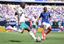 Soccer players in white-green and blue jerseys compete for a ball on a sunny field with a crowded stadium in the background.