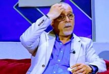 Man on a TV talk show sits on a red couch, hand on forehead, wearing a light blazer and glasses.