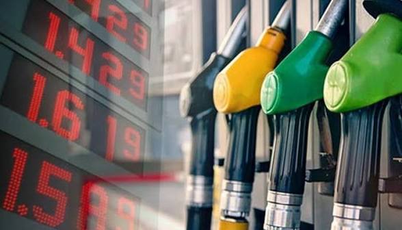 Close-up of multiple colored gas pump nozzles (yellow and green) at a pump, with a blurred red numerical price board in the background indicating fuel prices or market data.