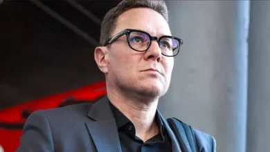 بنهاشم | MCG24 | موقع إلكتروني مغربي شامل متجدد على مدار الساعة Portrait of a man in a dark blazer and black shirt, wearing glasses, looking upward with a serious expression in a modern indoor setting.