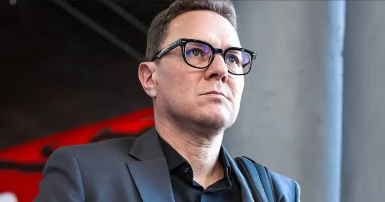 بنهاشم | MCG24 | موقع إلكتروني مغربي شامل متجدد على مدار الساعة Portrait of a man in a dark blazer and black shirt, wearing glasses, looking upward with a serious expression in a modern indoor setting.