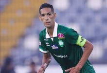 Soccer player in a green jersey with a yellow captain's armband, mid-match on the pitch.