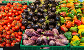 Colorful display of fresh vegetables at a market: tomatoes, purple eggplants, purple onions, and green and yellow bell peppers in crates.