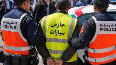 images 43 | MCG24 | موقع إلكتروني مغربي شامل متجدد على مدار الساعة Police detain a man in a crowd; two officers flank him, one in a bright pink-and-orange vest and another in navy, with a yellow vest reading 'حارس سيارات' (parking attendant).