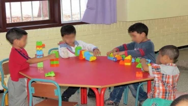 Four young children sit around a pink round table, stacking colorful plastic blocks in a classroom setting.