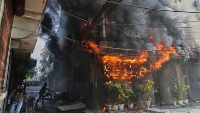 Storefront fire on a narrow street with thick black smoke and bright orange flames; bystanders nearby and tangled overhead wires above the scene.