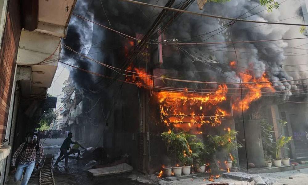 Storefront fire on a narrow street with thick black smoke and bright orange flames; bystanders nearby and tangled overhead wires above the scene.