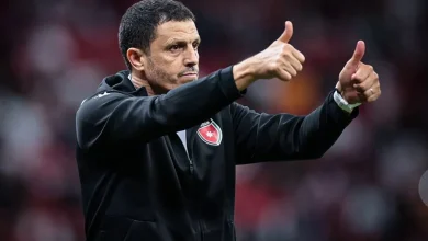 Coach on the sideline giving double thumbs up, wearing a black jacket with a team crest on the chest.