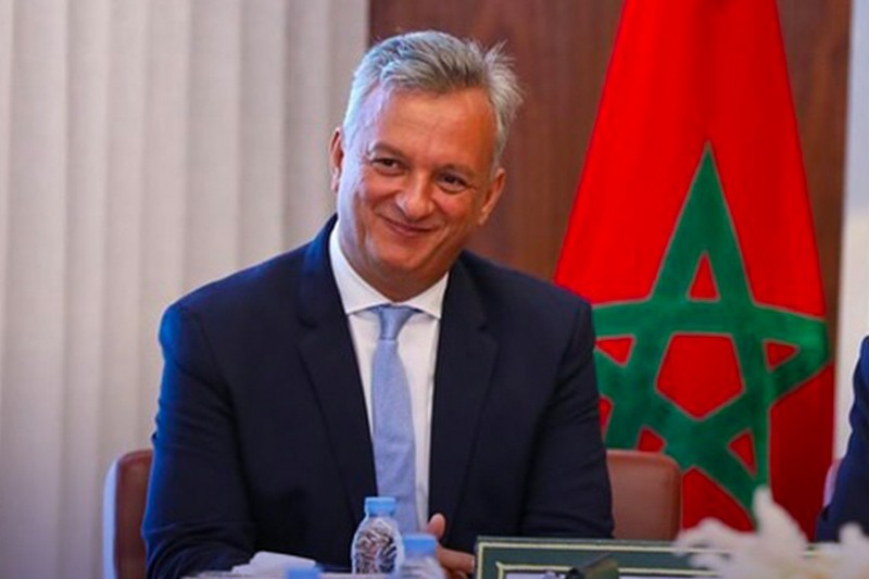 Smiling man in a dark suit and light blue tie seated at a table, Moroccan flag in the background.