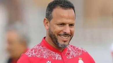 Smiling man in a red sports jersey with a white geometric pattern on the shoulders, outdoor blurred background.