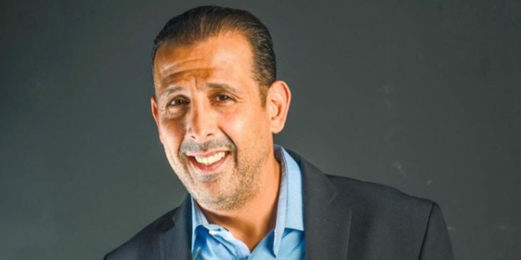 Portrait of a smiling man in a blue shirt and dark blazer against a dark gray backdrop.