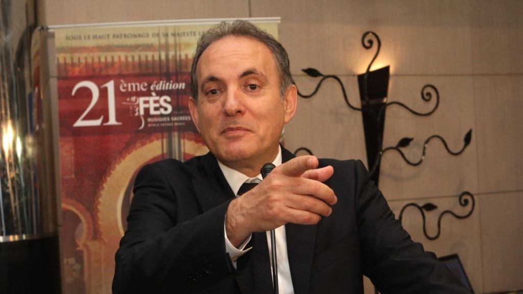 Man in a dark suit speaks at a podium, gesturing with his hands; a poster reading '21e édition FES' is visible in the background.
