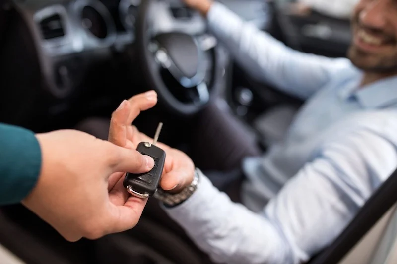 Close-up of one person handing a car key to a smiling driver seated inside a car.